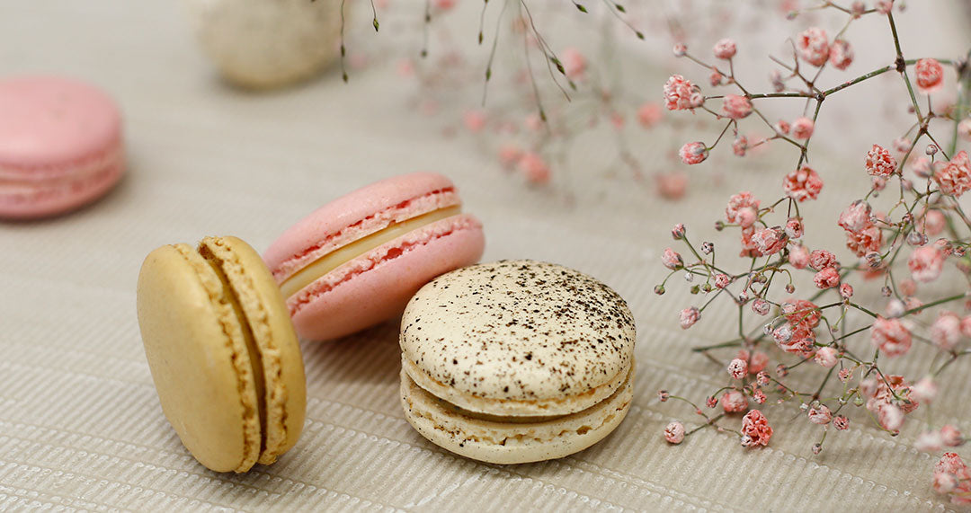 Three colorful macarons—pink, beige, and speckled white—surrounded by delicate pink flowers on a textured surface.